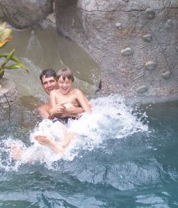 A father and son slide down in a swimming pool with a rock wall feature